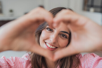 Naklejka premium Smiling young woman framing a heart with her hands, expressing love, warmth and connection. Casual indoor portrait conveying happiness, affection, friendliness and positive lifestyle mood.