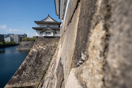 Osaka Castle rises above a peaceful Japanese garden with a reflective pond under a clear blue sky. A scenic travel destination in Osaka, Japan, during spring.