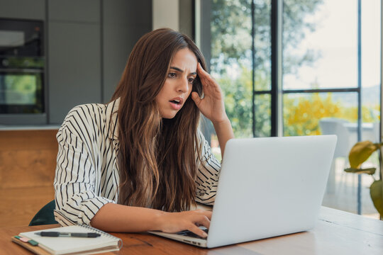 Stressed young woman working from home, looking shocked and frustrated at a laptop screen, holding her head as she encounters a significant problem or error