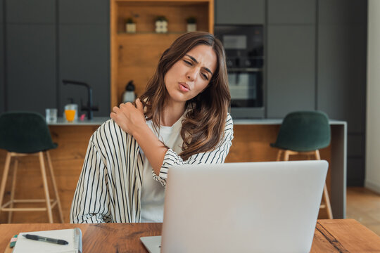 Woman feeling painful discomfort in her shoulder, rubbing a sore muscle while working from home on a laptop, experiencing stress and poor posture