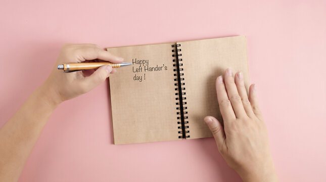 Left hand writing Happy Left Handers Day message in notebook on pink background