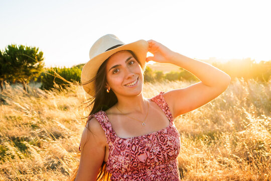 Young woman wearing a straw hat and patterned dress smiles in a sunlit field at golden hour, conveying summer warmth, carefree mood and natural beauty in a rural outdoor setting.
