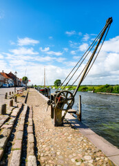 Fototapeta premium Historic harbor crane on Ribe's waterfront with sailing ships, river and traditional harbor buildings beneath a blue sky.