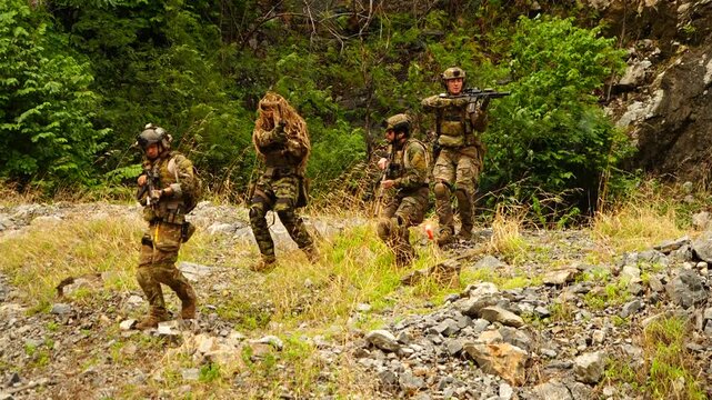 Five multinational soldiers in infantry uniforms, carrying paintball guns and ready for combat, run and practice military drills in a hilly, wooded area.
