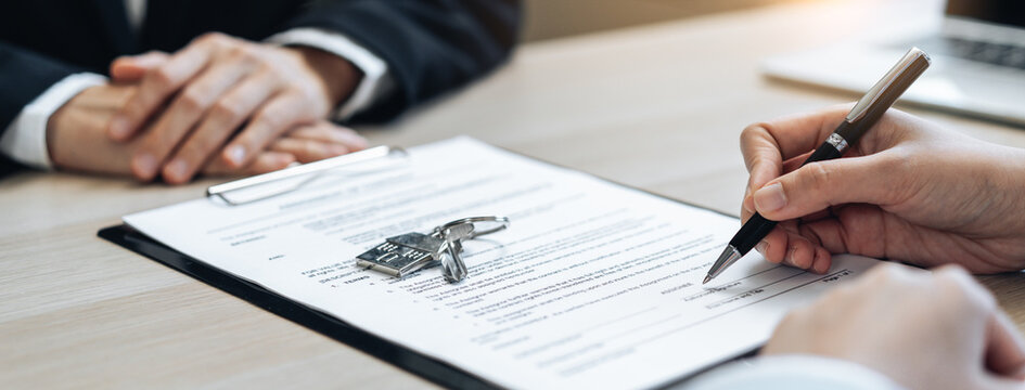 Close up of real estate client signing house purchase contract with keys on document.