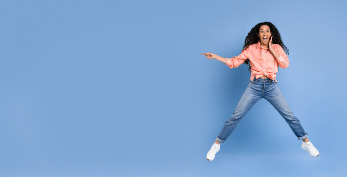 A woman is jumping in the air with a surprised expression on her face. She wears a pink shirt and blue jeans. The background is a solid blue color. This scene takes place during daytime