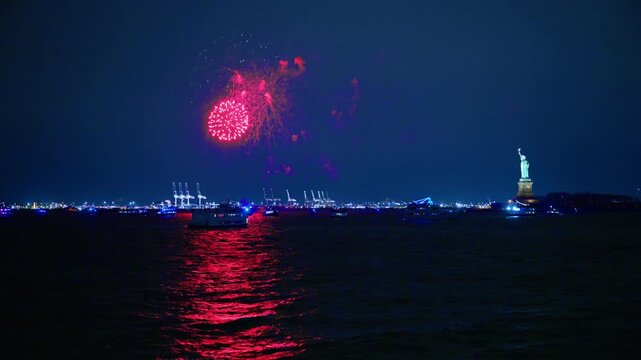 Fireworks exploding over New York Harbor near Statue of Liberty. Red burst reflected on water with boats, port cranes, and dark sky.
