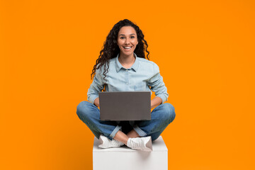 Fototapeta premium A woman with curly hair sits on a box with her legs crossed, using a laptop. She looks happy as she interacts with the device. The background is bright orange, creating a lively atmosphere