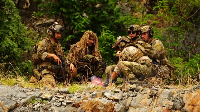 Five multinational soldiers in infantry uniforms, carrying paintball guns and ready for combat, hold a training exercise in a forested hillside.
