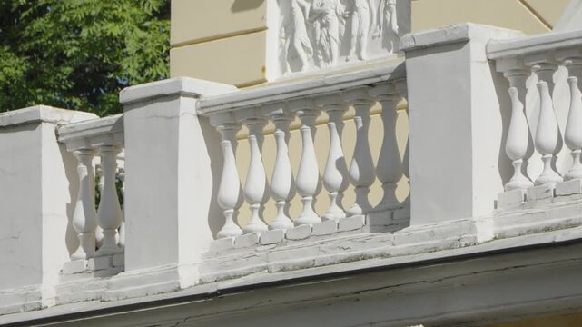 A bright, sunlit shot showcases the elegant white balustrade and bas-relief details of an old neoclassical building facade against a backdrop of green trees.