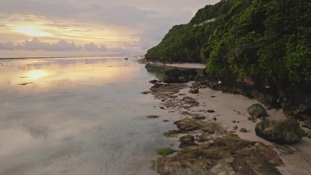 Low angle drone flight over ocean water mirrors a soft sunset sky beside a sandy beach and lush green cliff, creating a tranquil tropical coastline scene perfect for travel, relaxation, island escapes
