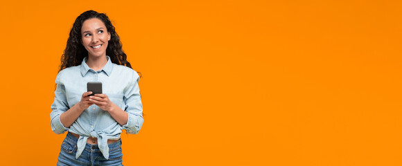 A woman stands with a smartphone in her hands and smiles. She is wearing a light shirt and jeans. The background is bright orange. The woman appears happy and engaged with her phone © Prostock-studio