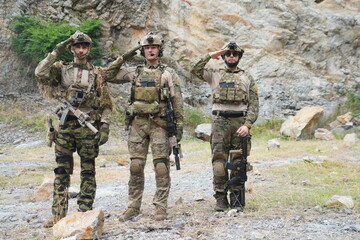 Fototapeta premium Three male soldiers in infantry uniforms are seen holding paintball guns and saluting during a military patrol training exercise in the forest. 
