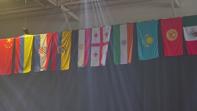 A panning shot displays a row of diverse national flags hanging indoors against a dark backdrop as light beams shine from above.
