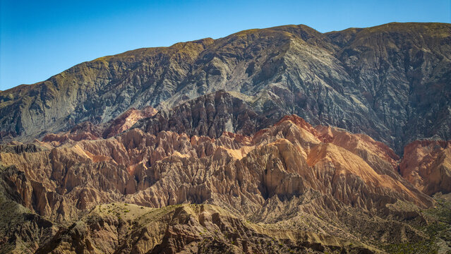 Aerial view of the colorful, rugged mountain ranges of the Andes with distinct geological layers and deep valleys under a clear blue sky in Salta, Salta Province, Argentina.