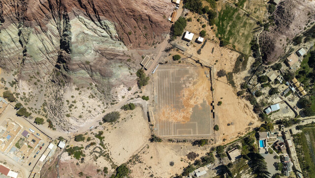 Aerial view of a dirt soccer field situated at the base of colorful sedimentary rock formations in the arid landscape of Salta, Salta Province, Argentina.