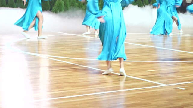 A cropped shot shows the legs and feet of multiple performers wearing elegant long blue dresses as they dance a synchronized routine on a wooden indoor basketball court floor with stage smoke.