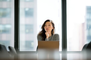 Fototapeta premium Asian woman sitting at office desk with laptop thinking about business strategy. Focused employee looking away and planning project. Corporate work, professional career and ambition.