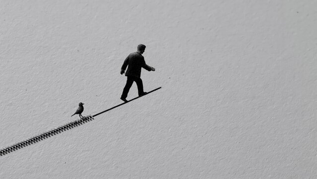 Miniature man and bird walk along thick metal tightrope.