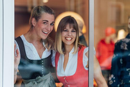 Two smiling women in authentic Bavarian dirndl outfits admire a traditional clothing shop window celebrating local culture and heritage.