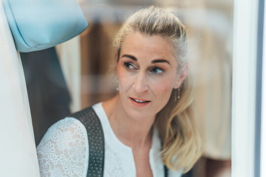 Elegant blonde woman wearing a Bavarian dirndl standing behind the shop window of a traditional Tracht store with a timeless portrait.