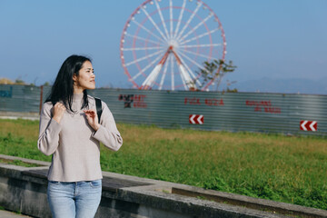 Obraz premium Woman with backpack strolling along a park path, with a large ferris wheel in the background, casual outfit under bright daylight, outdoor leisure and travel mood