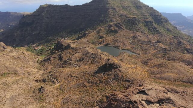 Stunning aerial perspective flying over the rugged and arid mountains of Mogan, revealing a winding road and a small dam in Gran Canaria