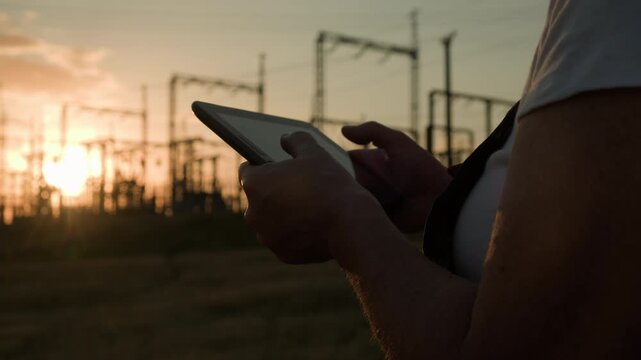 Electrical engineer's hands typing on tablet against background of high voltage transformer substation in evening at sunset