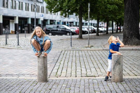 Brother sister spend playful time together city street while climbing concrete bollards. Authentic sibling relationship captures childhood, family bonding, curiosity, independence urban adventure