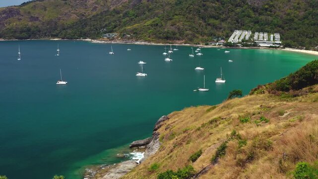 Aerial view showing multiple sailboats and yachts anchored in the turquoise water of Nai Harn Beach bay, surrounded by lush green hills and a sandy shoreline under a bright sky in Phuket, Thailand