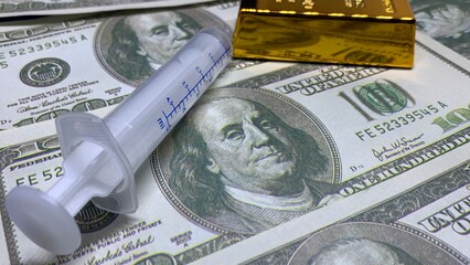 Plastic medical syringe placed on U.S. hundred-dollar bills with gold bar, representing healthcare expenses and financial value © MR HEPPY