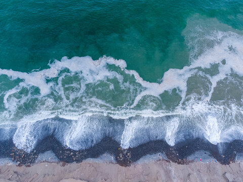 Aerial view of Pacific Ocean waves crashing onto the dark sandy beach with white sea foam patterns in Lima, Callao Region, Peru.