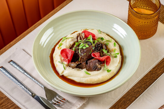 Delicious meal served in a bowl with meat and sauce on mashed potatoes. A dish features meat on top of mashed potatoes with sauce and fresh greens.