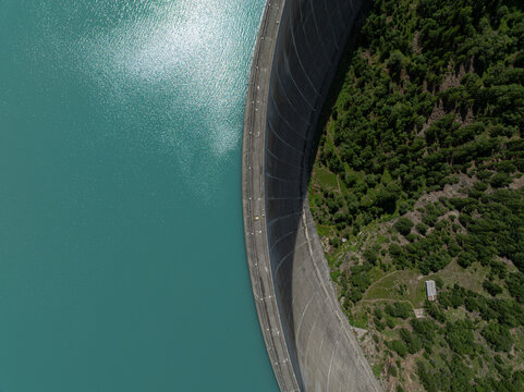 Aerial view of the curved concrete wall of the Place-Moulin Dam holding back turquoise reservoir water, surrounded by dense alpine forests in Bionaz, Aosta Valley, Italy.