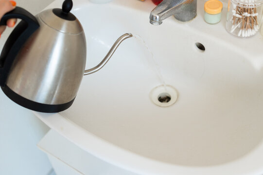 Hot water is poured from a stainless steel electric kettle into a white ceramic sink drain. The process is used for cleaning and unclogging the bathroom basin pipe.