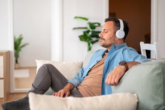 Mature man relaxing at apartment, enjoying sound with wireless headphones, finding peace and comfort by closing eyes and meditating in a tranquil environment