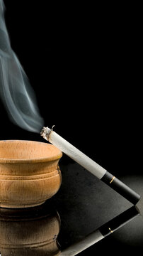 A cigarette burns slowly in a wooden ashtray while smoke curls up into the air in a dimly lit setting. The scene captures a quiet moment of smoking indoors.