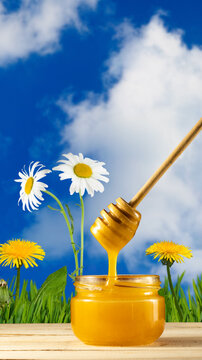 Honey is dripping from a wooden dipper into a jar on a table. Bright yellow flowers and green grass are in the background under a blue sky with clouds.