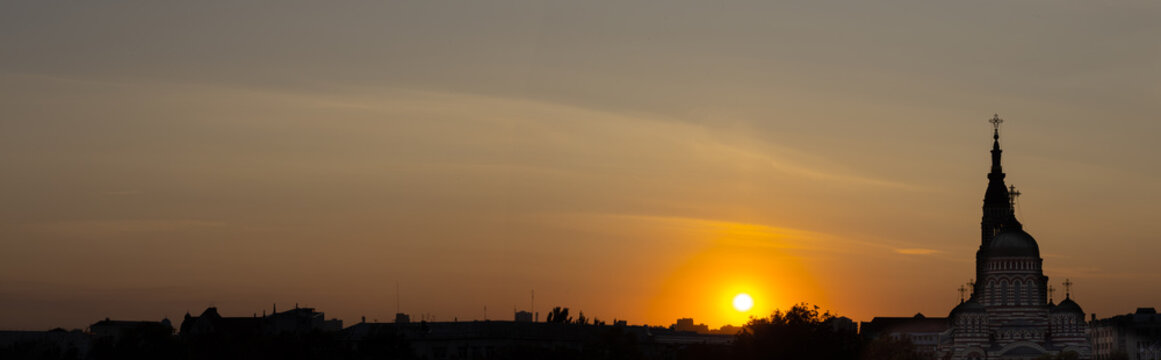 The sun sets behind a city skyline. A church tower stands tall against the colorful sky. The scene shows the end of the day with fading light.