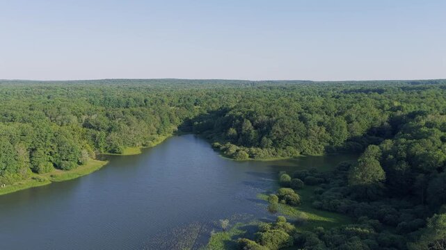 Aerial footage showcasing Lac du Bourdon in France, featuring a tranquil lake bordered by dense green forest and open meadows under a clear blue sky. The video transitions from views of the water