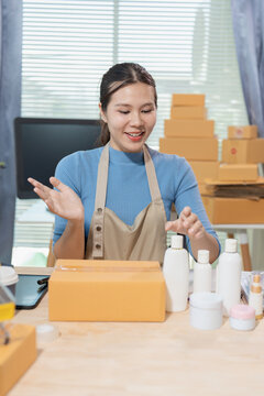 Smiling young Asian woman online seller packaging cosmetics skincare products into cardboard parcel boxes at home office, E-commerce small business owner concept.