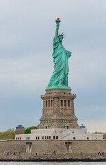 Fototapeta premium Statue of Liberty with full pedestal and visitors on Liberty Island, New York. Green copper figure on granite base and star fort, small figures of tourists on promenade, overcast sky.