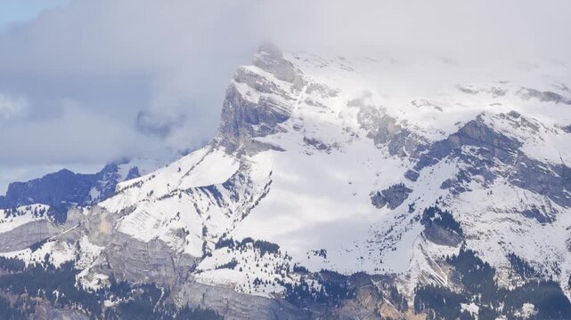 A dramatic close-up view of a rugged snow-covered mountain peak in the Alps, partially shrouded by clouds. The scene captures the steep rocky slopes, patches of evergreen forest, and the thick