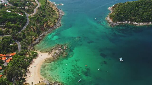 Tropical island landscape, turquoise sea water bordering Nai Harn Beach, Phuket. With winding coastal road and lush green hills leading to beachfront houses under a clear sky. Aerial view drone flight