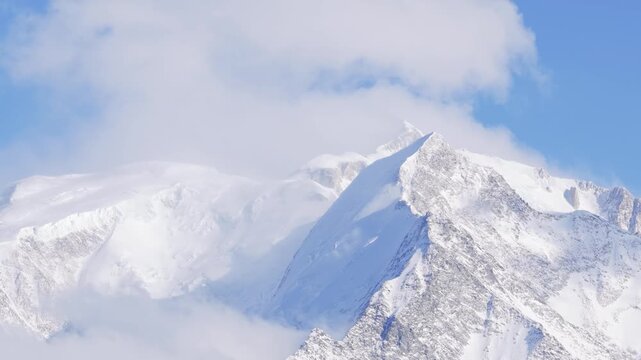 Cinematic close-up video of the snow-covered peaks of the Mont Blanc Massif, with dramatic clouds drifting across the mountains under a clear blue sky. The footage captures the pristine winter