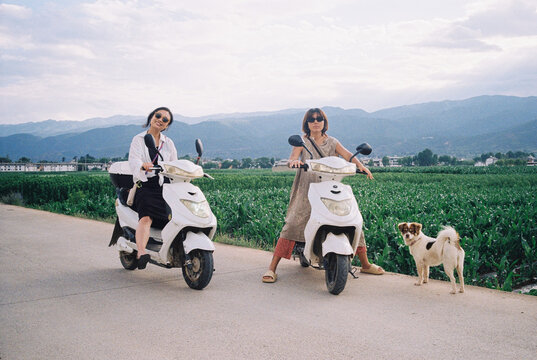 Two Women on Scooters by a Field With Mountains and a Dog