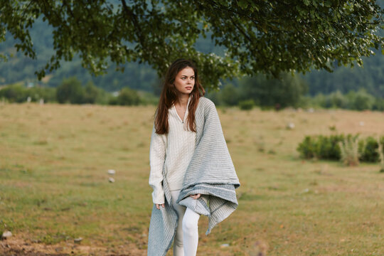A woman in a soft shawl stands in an open field, distant hills behind, looking away with a contemplative expression. A quiet, lifestyle moment in outdoor fashion.