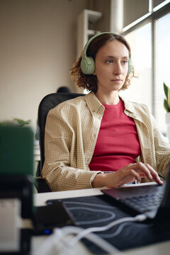 Vertical shot of young adult Caucasian woman wearing headphones and typing on laptop at desk, focusing on office work. Ideal for productivity, office work, programming