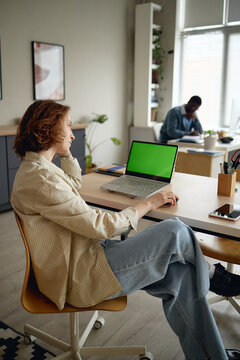 Young adult Caucasian woman working at laptop with green screen in office, multitasking at desk while colleague working in background. Useful for workplace technology or mockup marketing