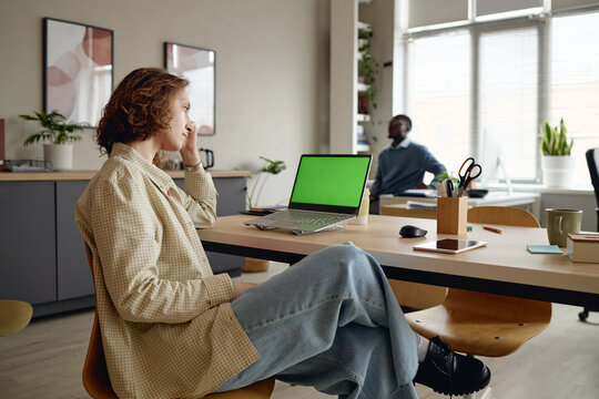 Young adult Caucasian woman sitting at office desk, touching face and thinking beside laptop with green screen. Ideal for workplace stress, burnout, productivity, mockup advertising